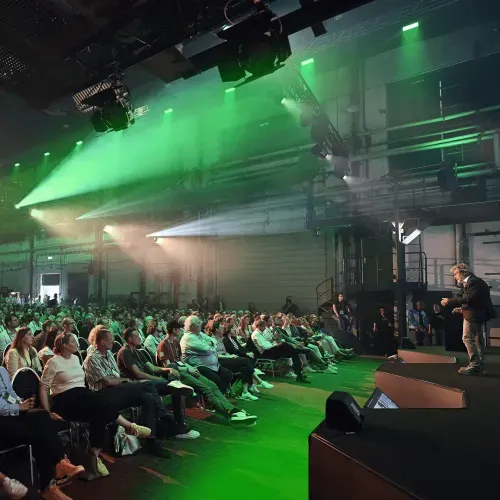 Speaker on stage presenting to a large audience seated in an industrial venue with green and white stage lighting.