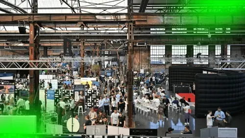 Crowded indoor expo hall with various booths, attendees walking and sitting in rows of chairs under industrial beams.