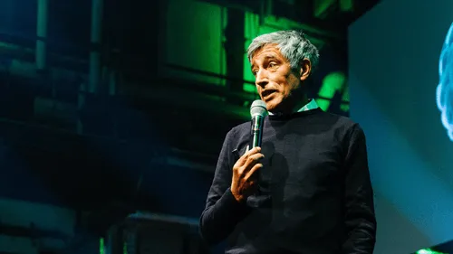Man with gray hair speaking into a microphone on stage with green lighting in the background.