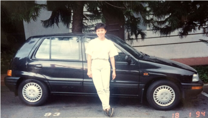 A woman with white T shirt smiling warmly in front of a car.