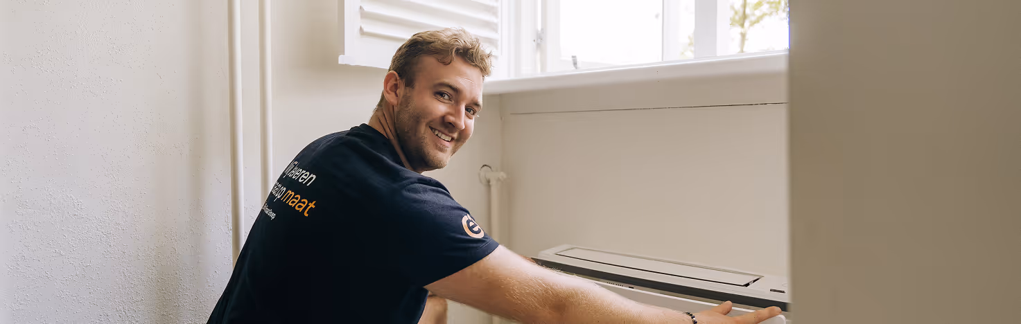 Monteur van Elite Klimaat Groep kijkt tevreden omhoog terwijl hij een aircosysteem installeert bij een open raam.
