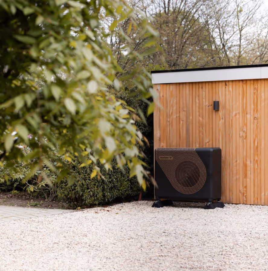 Zwarte warmtepomp buiten geplaatst naast een houten schuur in een groene tuin