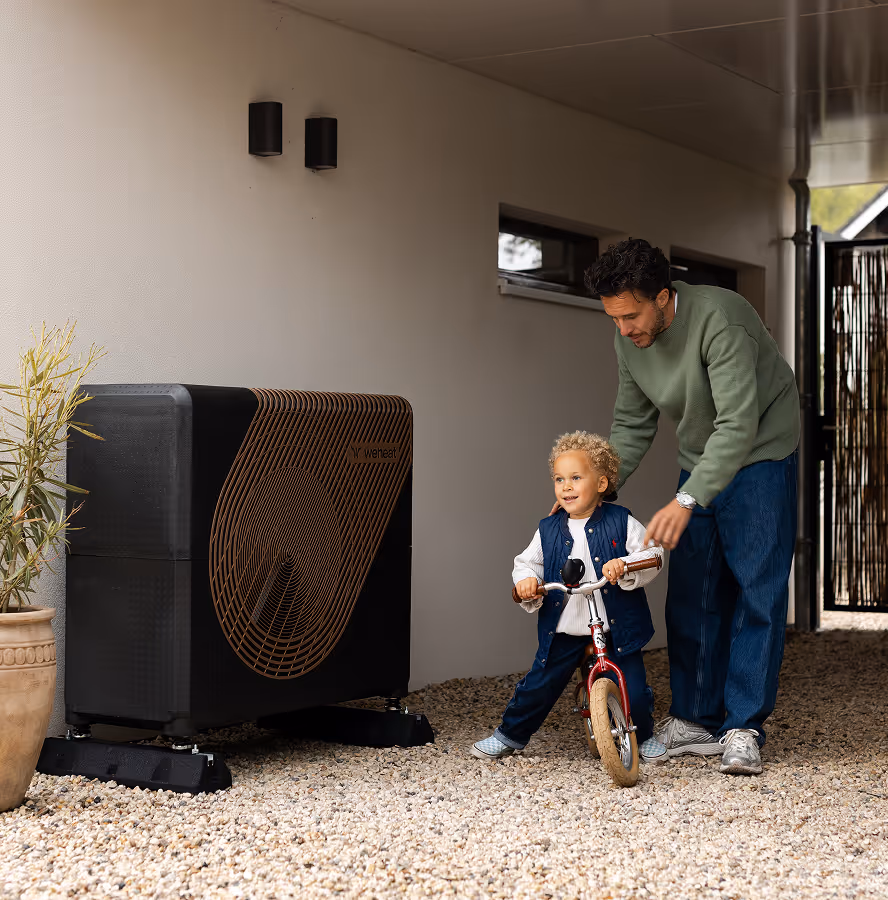 Vader helpt kind op loopfiets naast een zwarte warmtepomp onder een overkapping