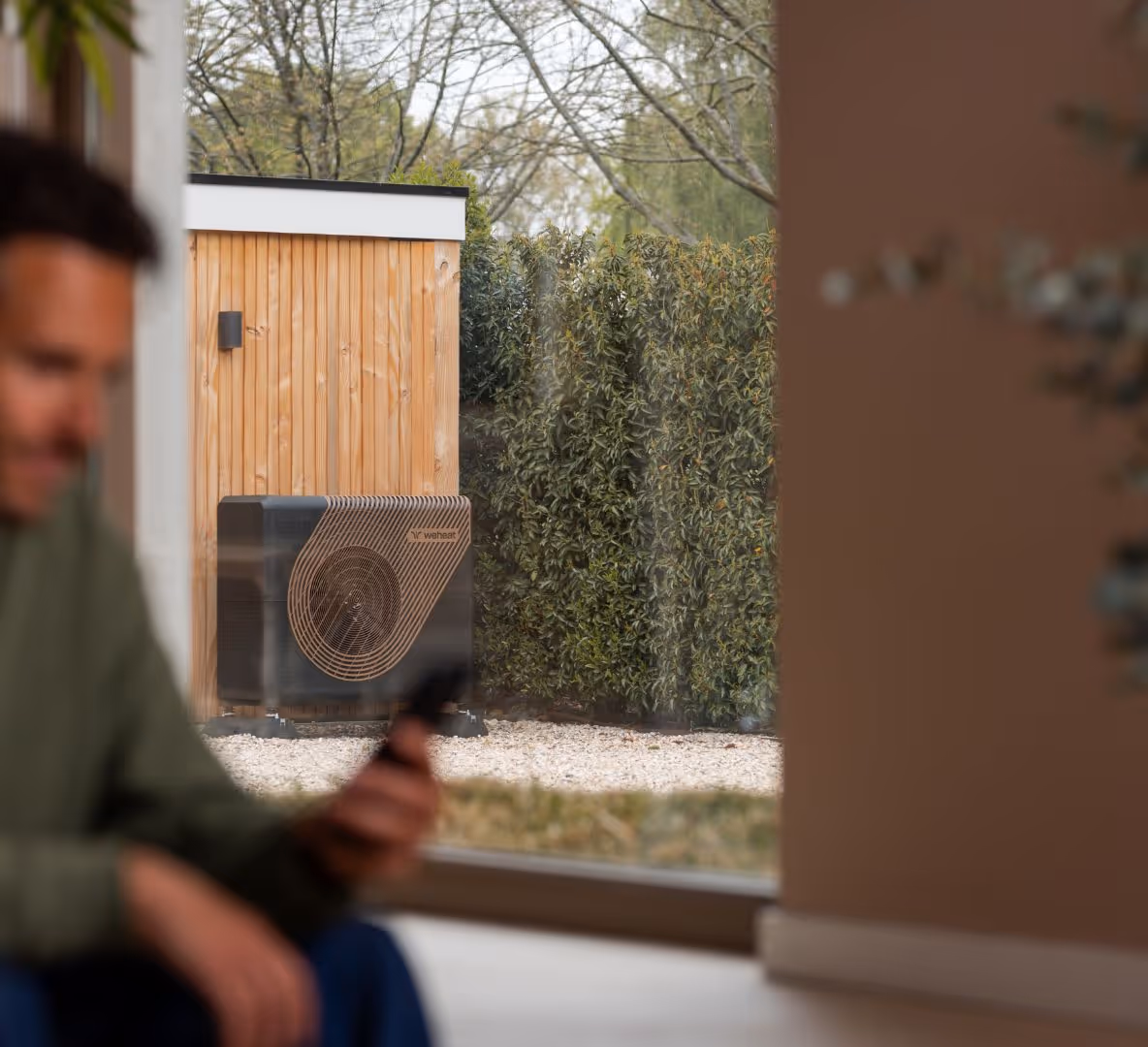 Buitenunit van een warmtepomp in de tuin, gezien door een raam vanuit de woning.