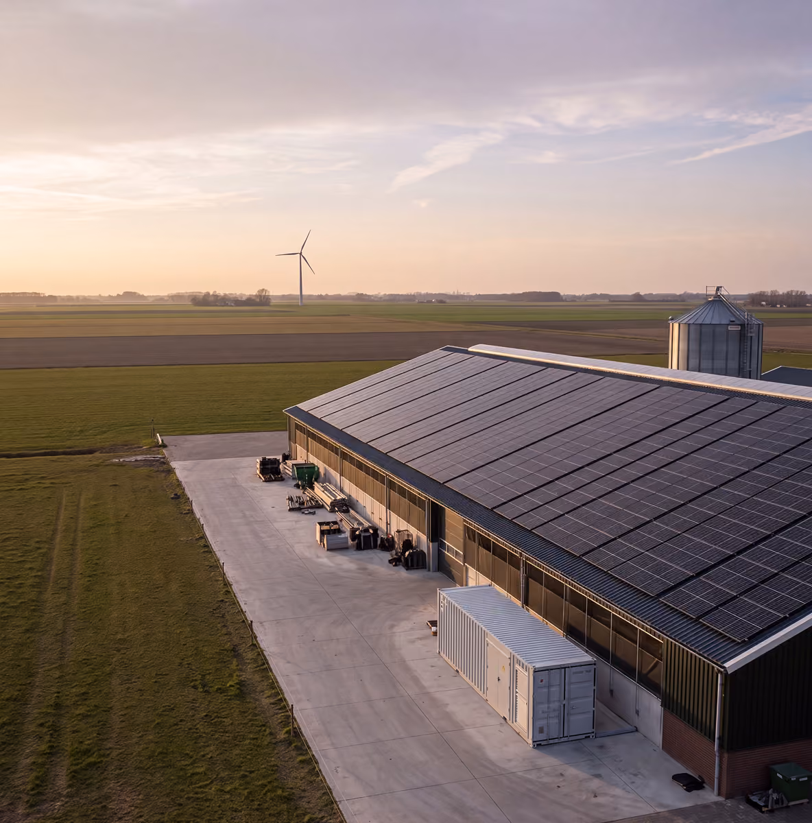 Luchtfoto van een agrarische loods met zonnepanelen op het dak en een batterijcontainer, met windmolen en akkers op de achtergrond