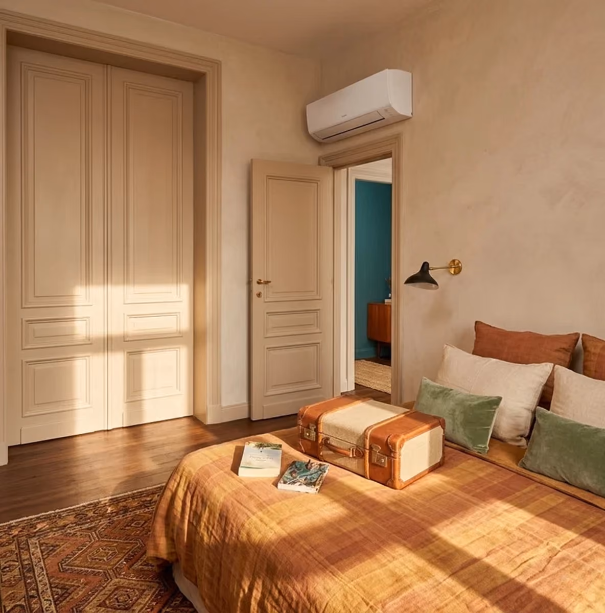 Daikin wandmodel airco boven de deur in een warme slaapkamer met bed, kussens en een vintage koffer