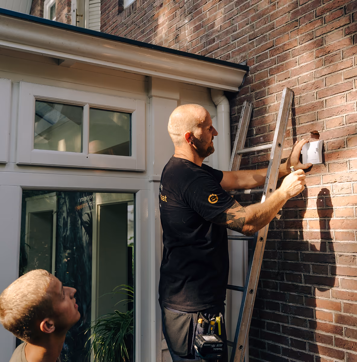 Monteur van Elite Klimaat Groep op een ladder monteert een muurdoorvoer voor een airco op een bakstenen gevel, collega kijkt toe