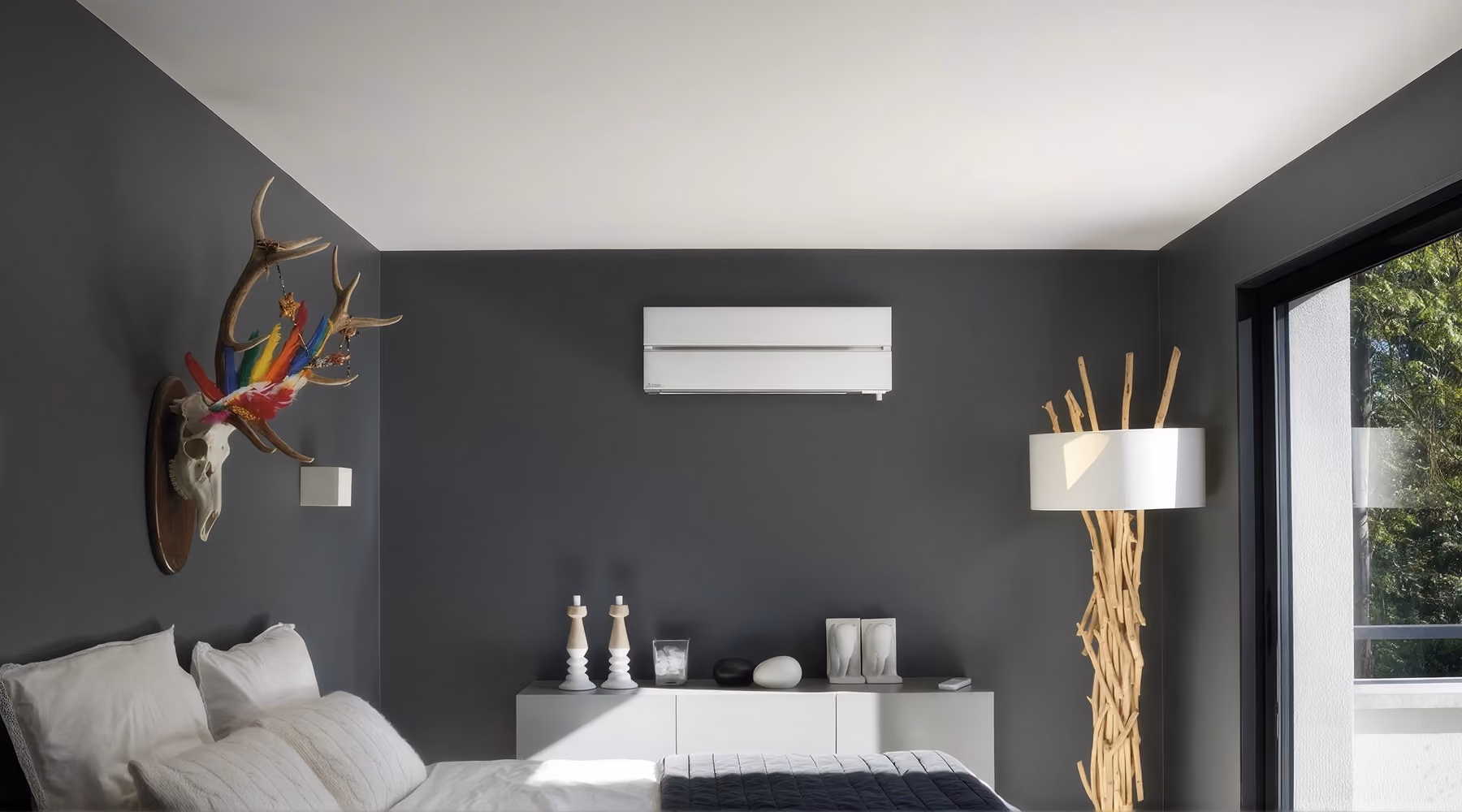 Witte Mitsubishi wandmodel airco op een antracietgrijze wand in een slaapkamer met wit dressoir en houten vloerlamp
