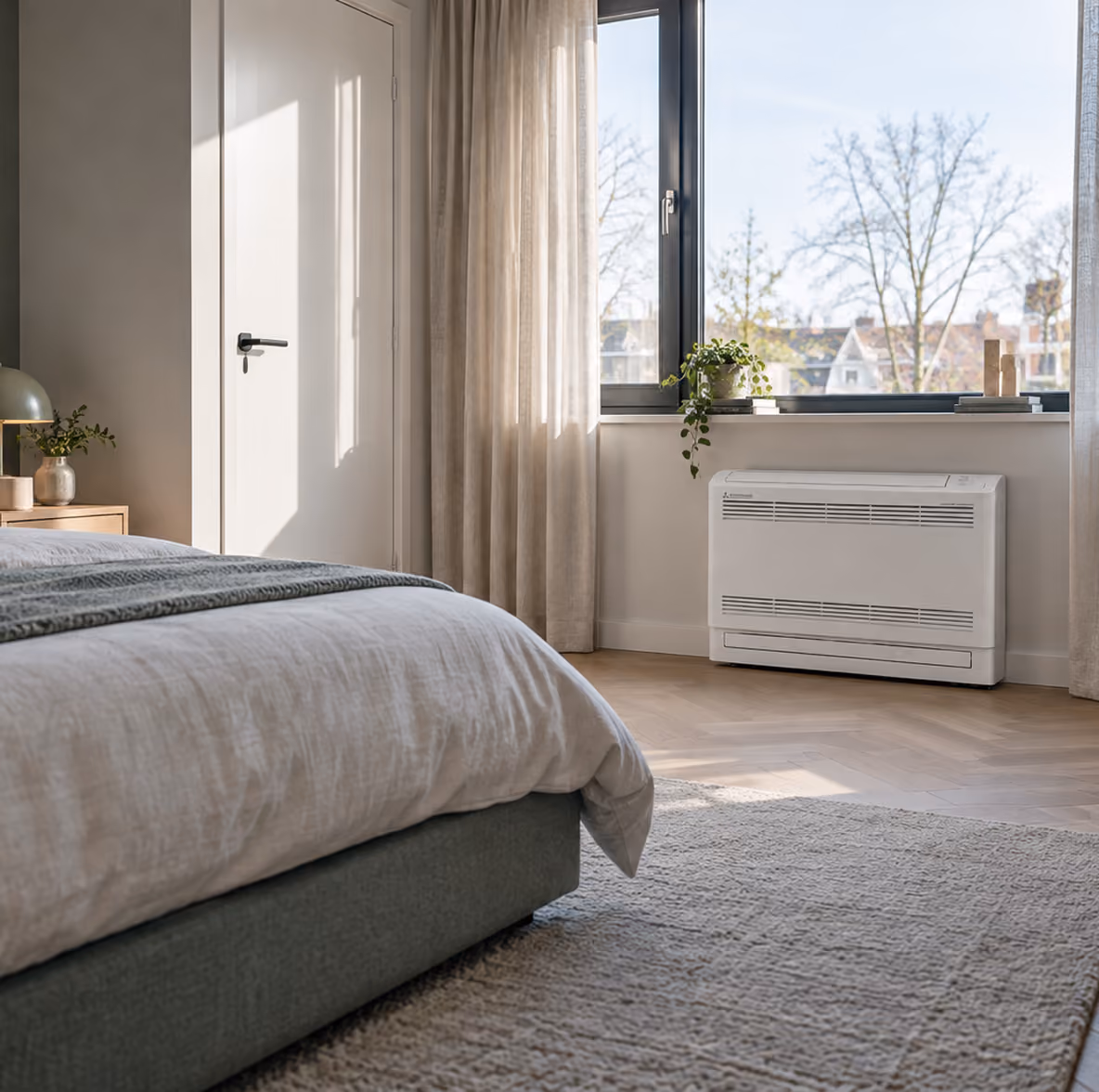 Mitsubishi vloermodel airco onder het raam in een zonnige slaapkamer met visgraatvloer en linnen gordijnen
