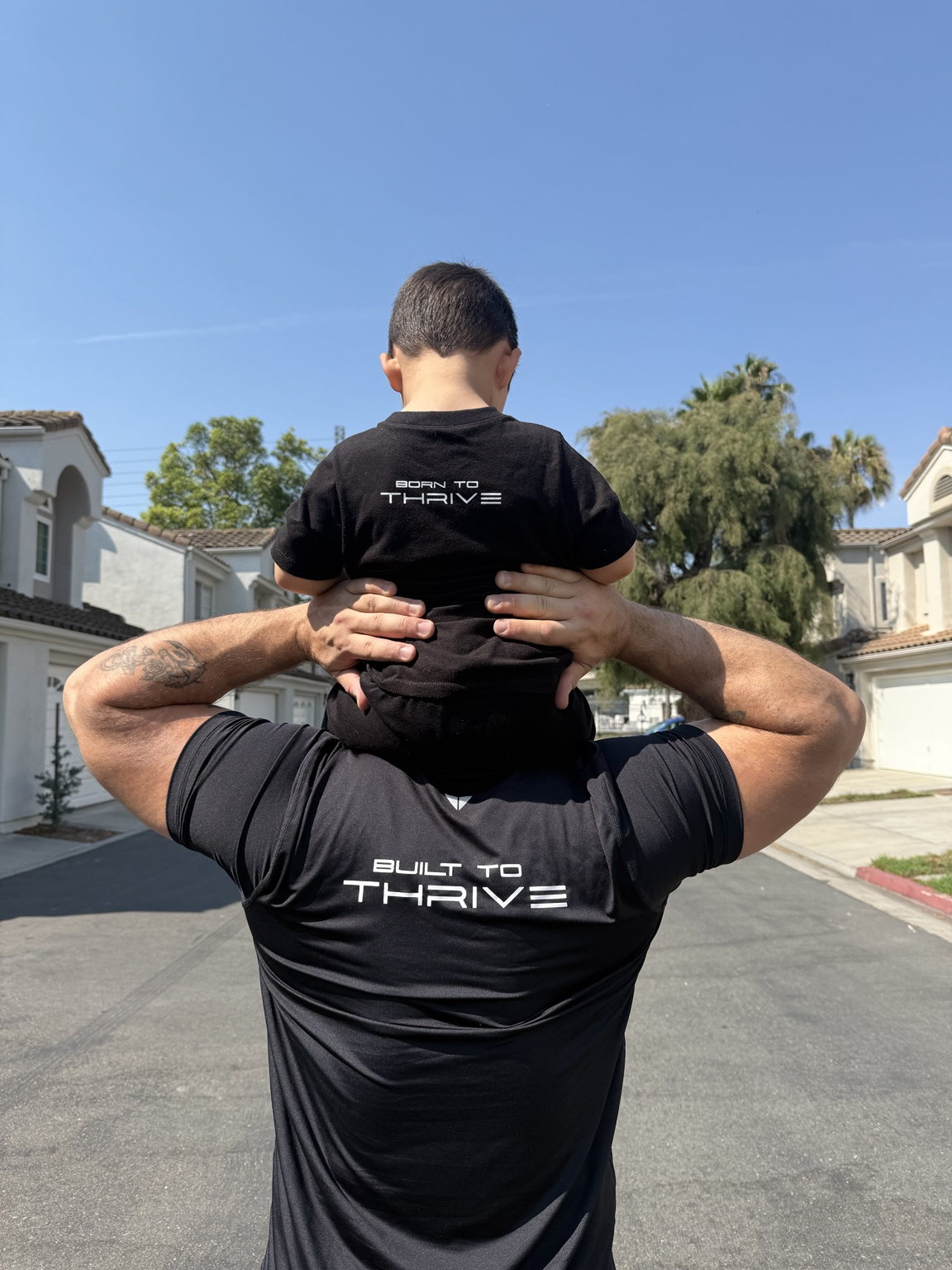 Joe El Khechef carrying his son Dany on his shoulders, wearing Built to Thrive and Born to Thrive shirts.