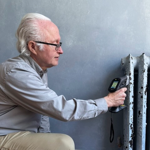 Home inspector with white hair and glasses measuring temperature of a radiator with a handheld device against a gray wall.