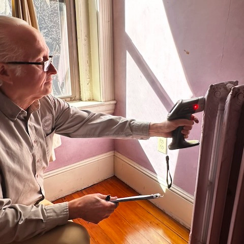Jack Mason inspects a purple wall near a window using a handheld infrared thermometer and a metal tool.