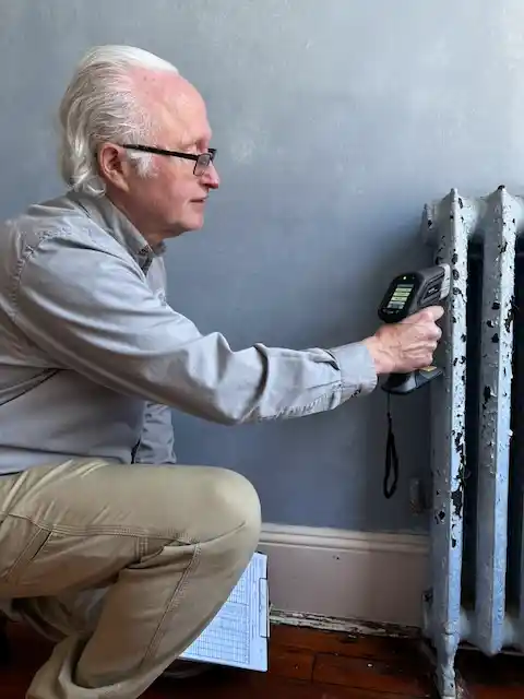 Jack Mason kneeling indoors scanning an old metal radiator with a handheld electronic device.