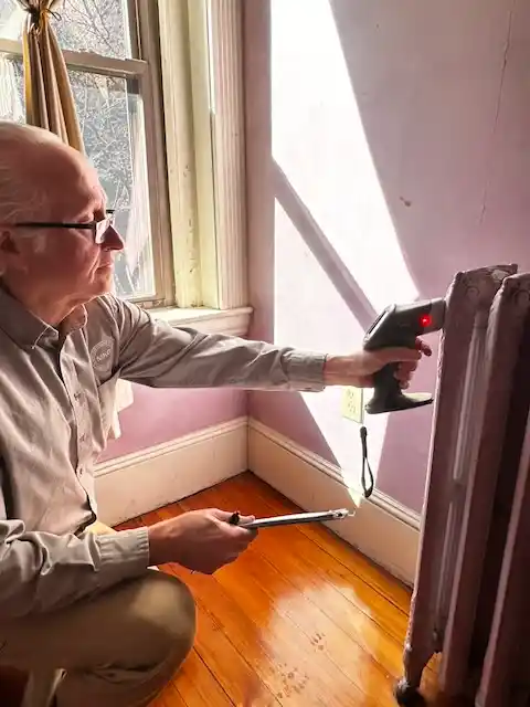 Jack Mason kneeling near a window using a handheld infrared thermometer on a radiator.
