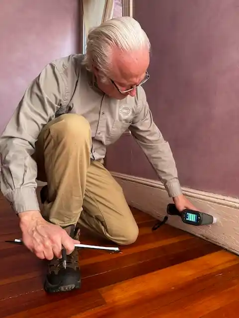 Jack Mason kneeling on wooden floor, using a handheld electronic device to inspect the baseboard.