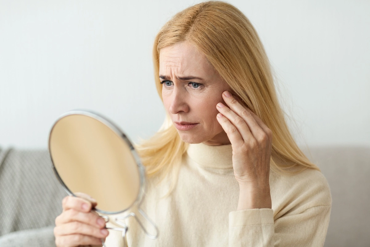Middle-aged woman with concerned expression looking in a mirror and touching wrinkles on her face