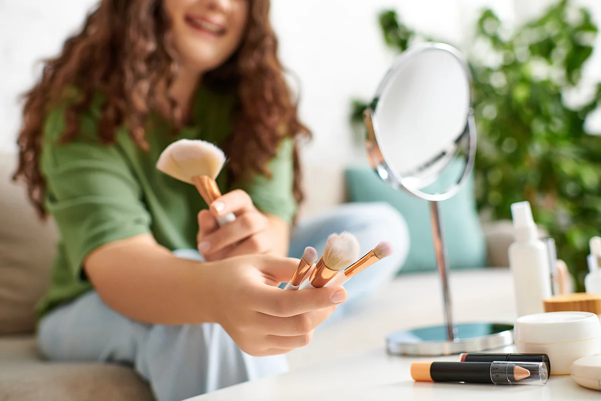 Woman holds beauty blender and brushes in home setting.