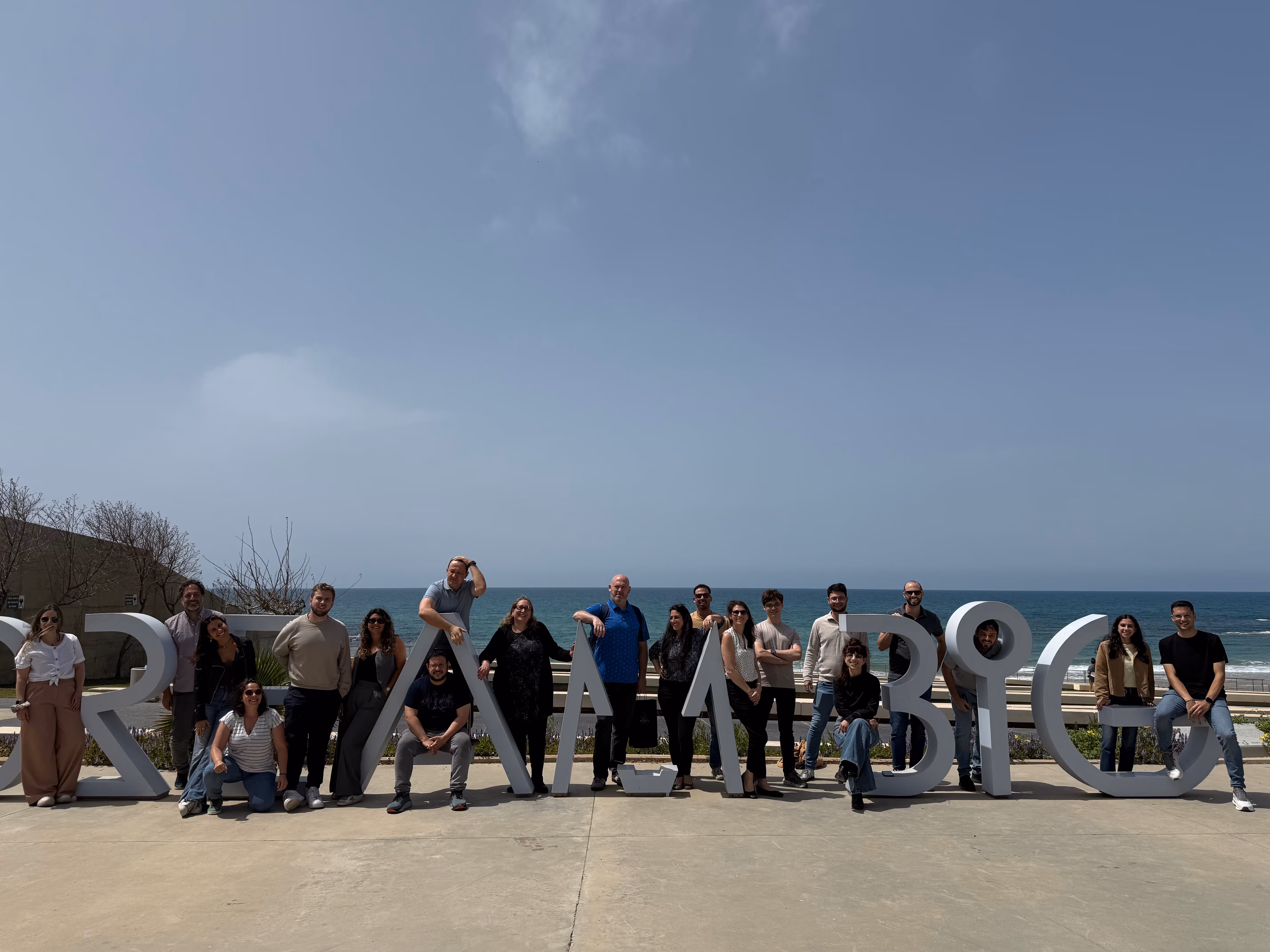 Group of people posing with large geometric sculpture letters by the seaside under a clear sky.
