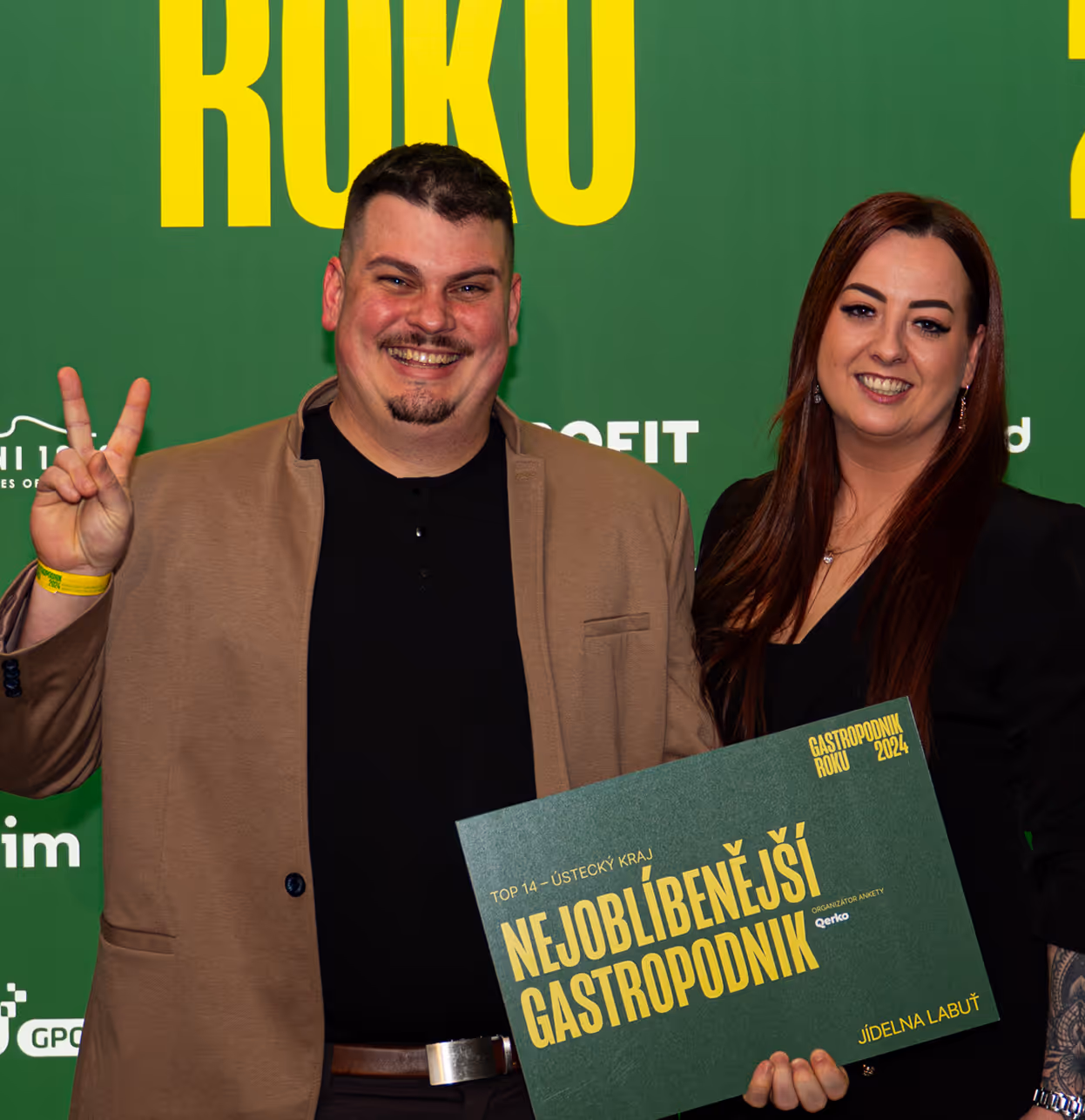 Smiling man in a brown jacket making a peace sign and woman holding a green award sign with yellow text in front of a green backdrop.
