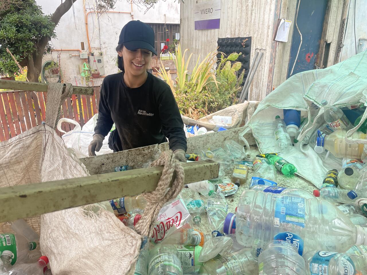 Smiling worker wearing a black cap and gloves sorts plastic bottles at an outdoor recycling facility, with large white sacks filled with collected plastics in the background.