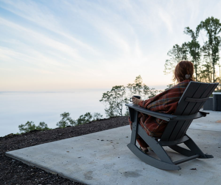 A young woman sitting in a rocking chair on the patio of her prefab mountain cabin. 