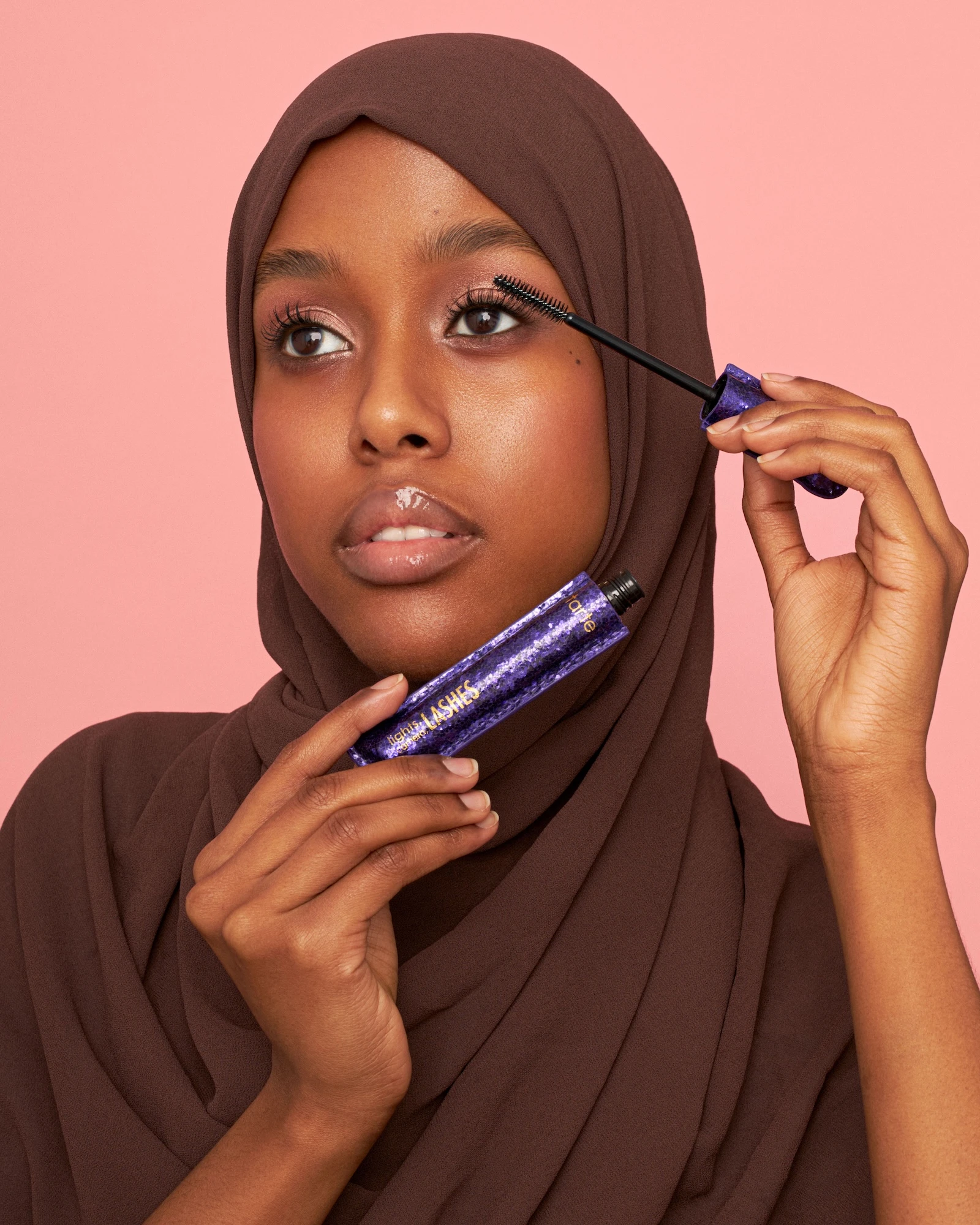 A woman with a brown head scarf holding a black and purple makeup brush.