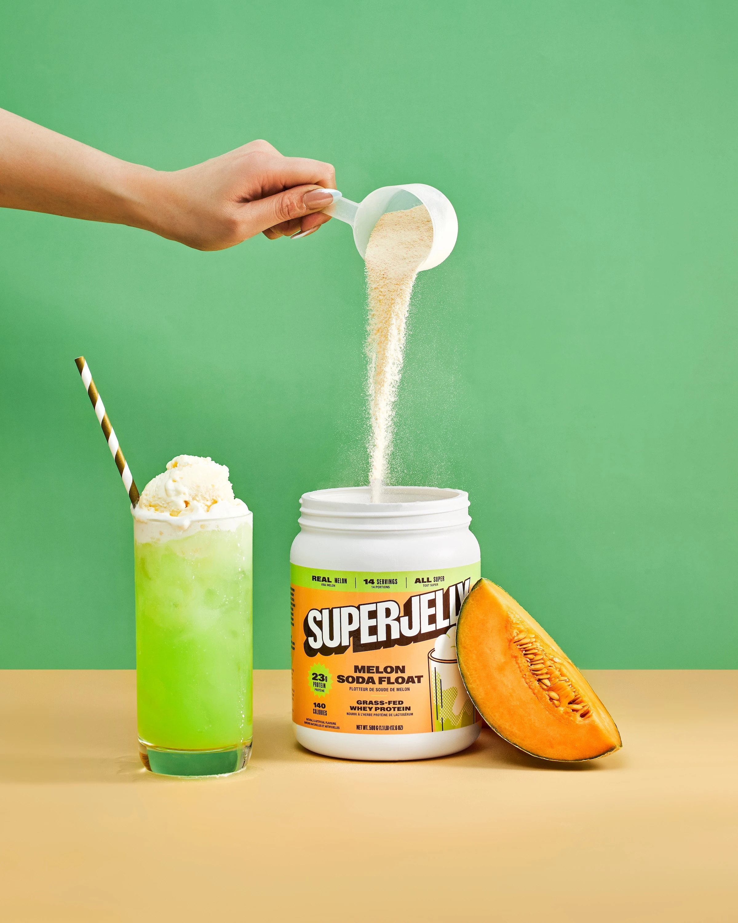 A person pouring powder into a container with a green drink and a peeled orange.