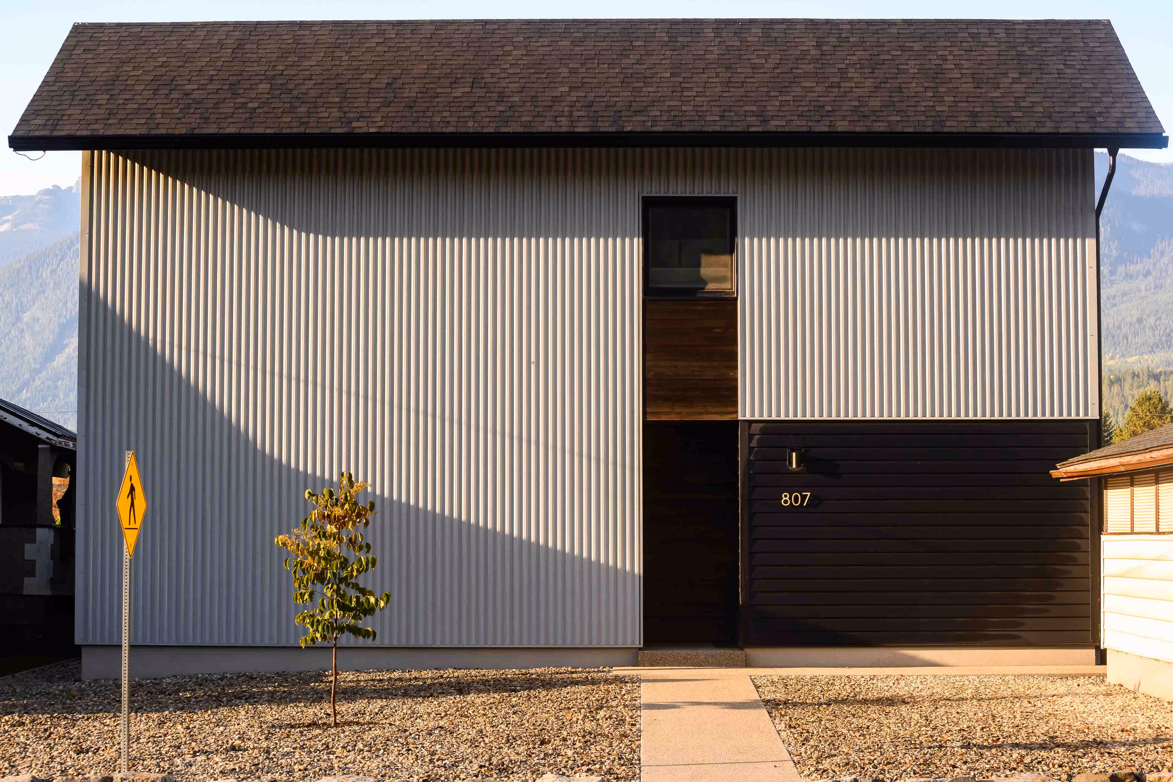 Modern house with gray vertical siding, a dark entrance, and the number 807 on the right side, with a small tree and pedestrian crossing sign in front.