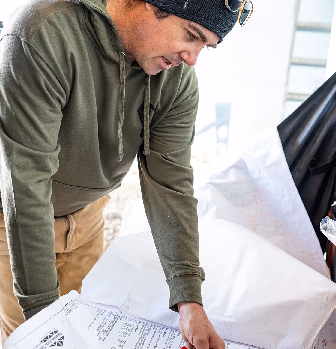 Man wearing a green hoodie and black beanie leaning over a table reviewing architectural or construction blueprints.