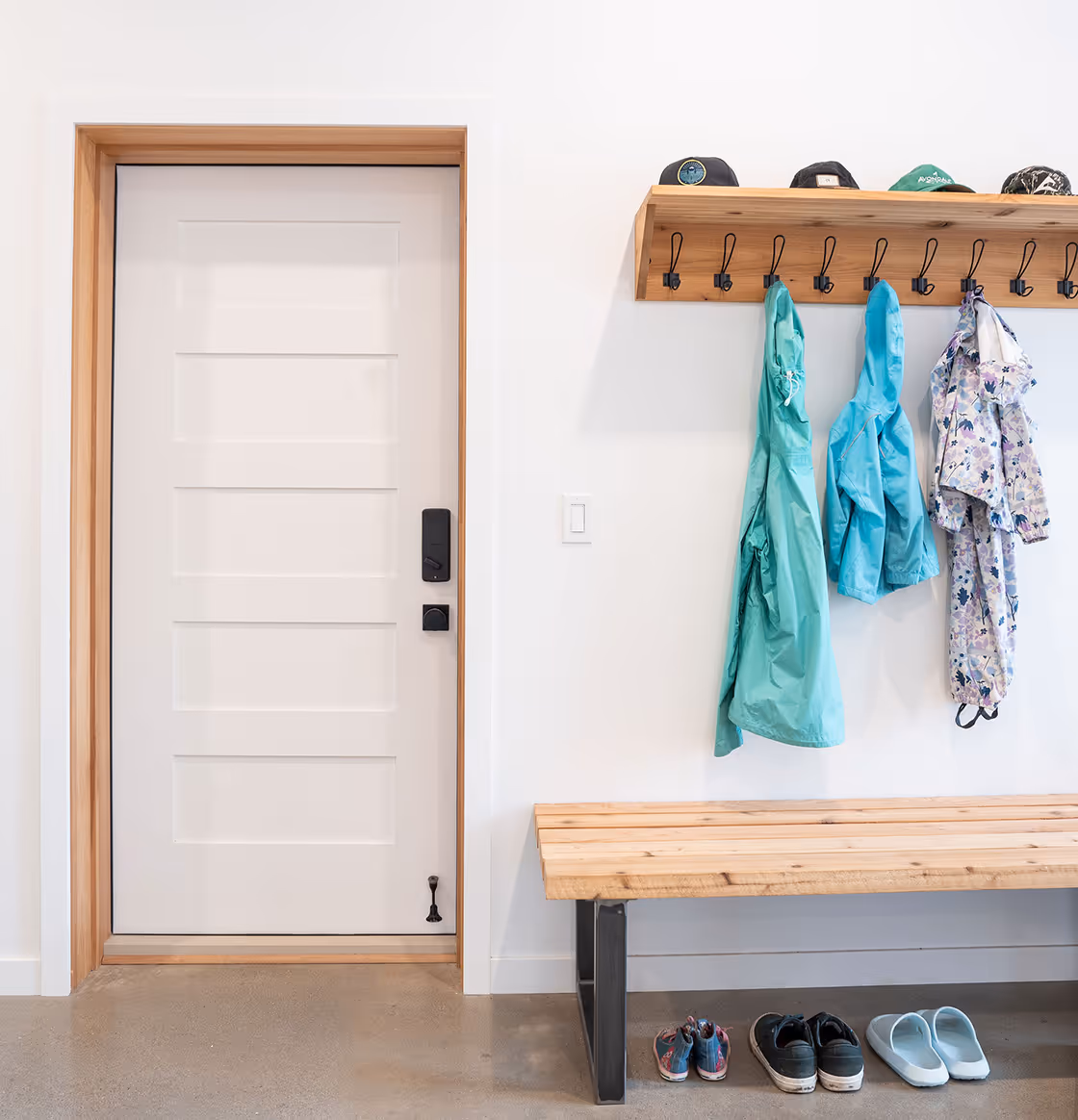 Clean, modern entryway with a closed white door, wooden bench, wall-mounted coat rack holding rain jackets, and shoes neatly placed below.