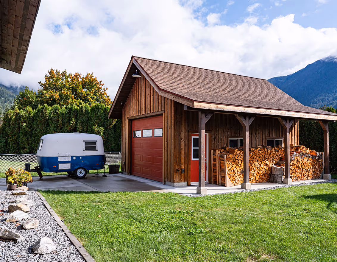 Wooden garage with a red door and stacked firewood under a side overhang, next to a small blue and white camper trailer on a concrete driveway surrounded by green grass and trees.