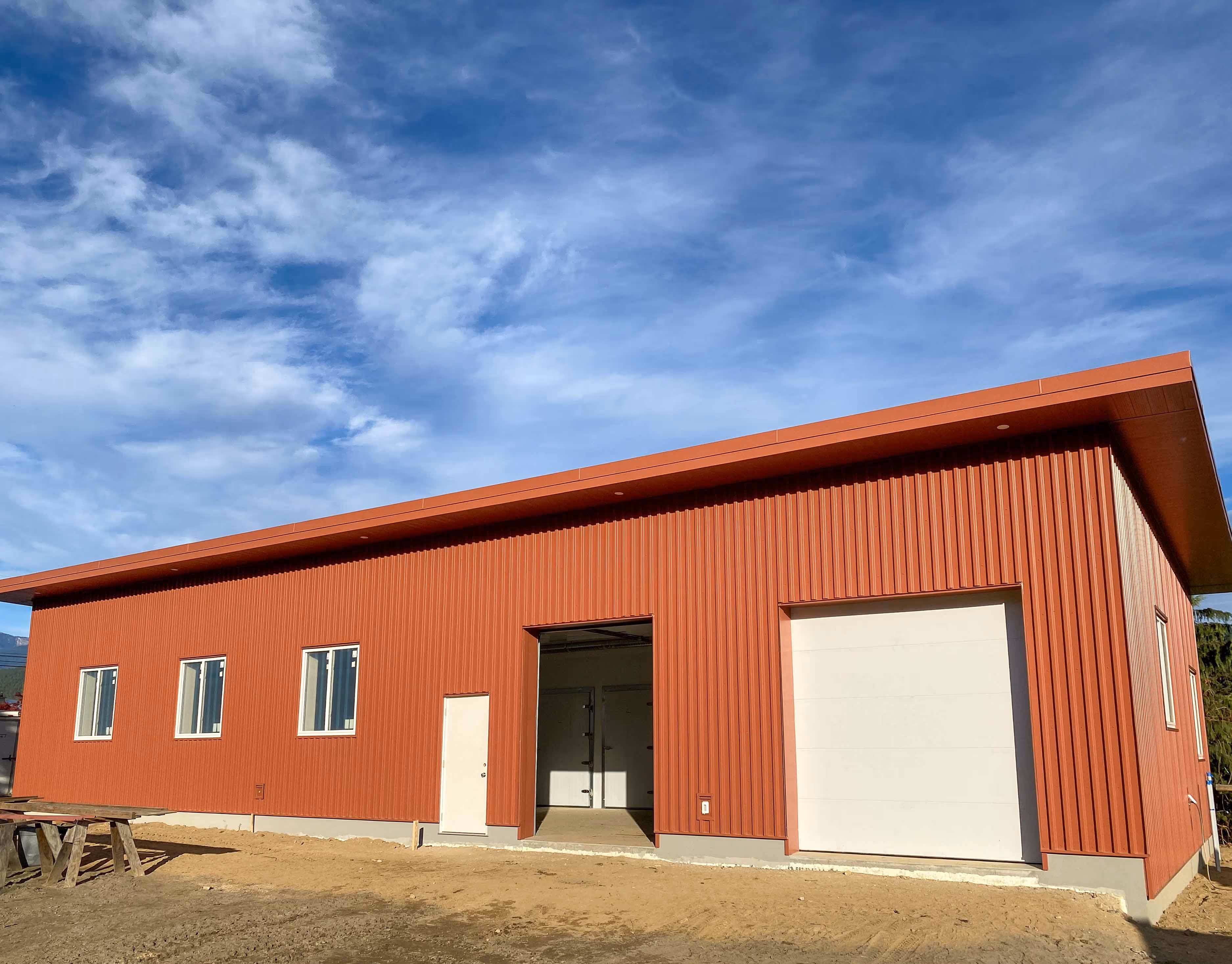 Large red metal commercial building with three windows, a regular door, an open doorway, and a closed white garage door under a blue sky with clouds.