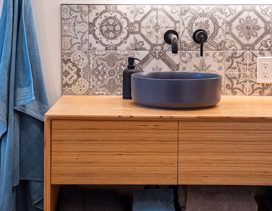 Modern bathroom sink with matte black faucet and soap dispenser on a wooden vanity, patterned gray and white tile backsplash, and blue towel hanging nearby.