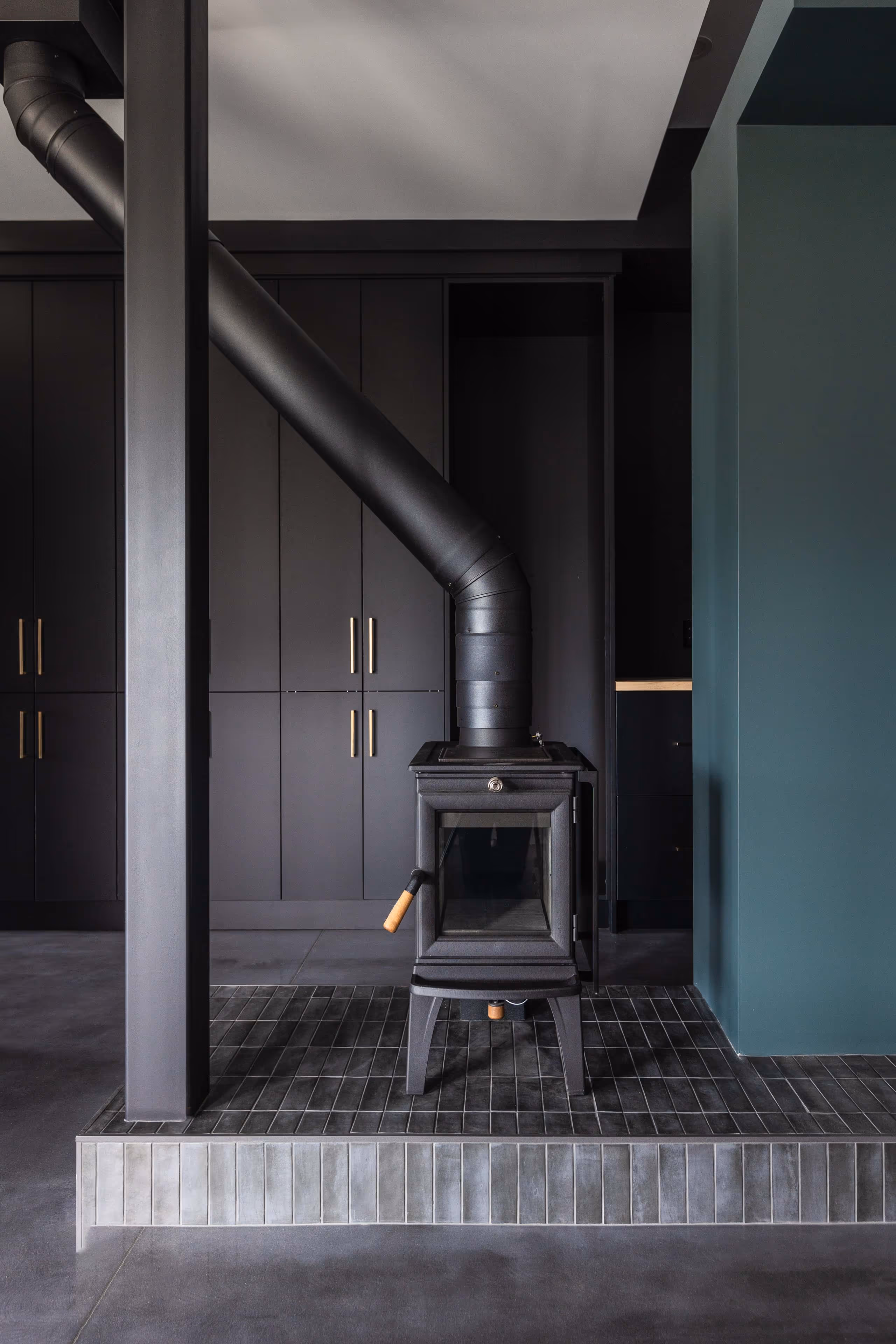 Black wood-burning stove with angled chimney pipe on dark tiled platform in a modern interior with dark cabinetry and teal wall.