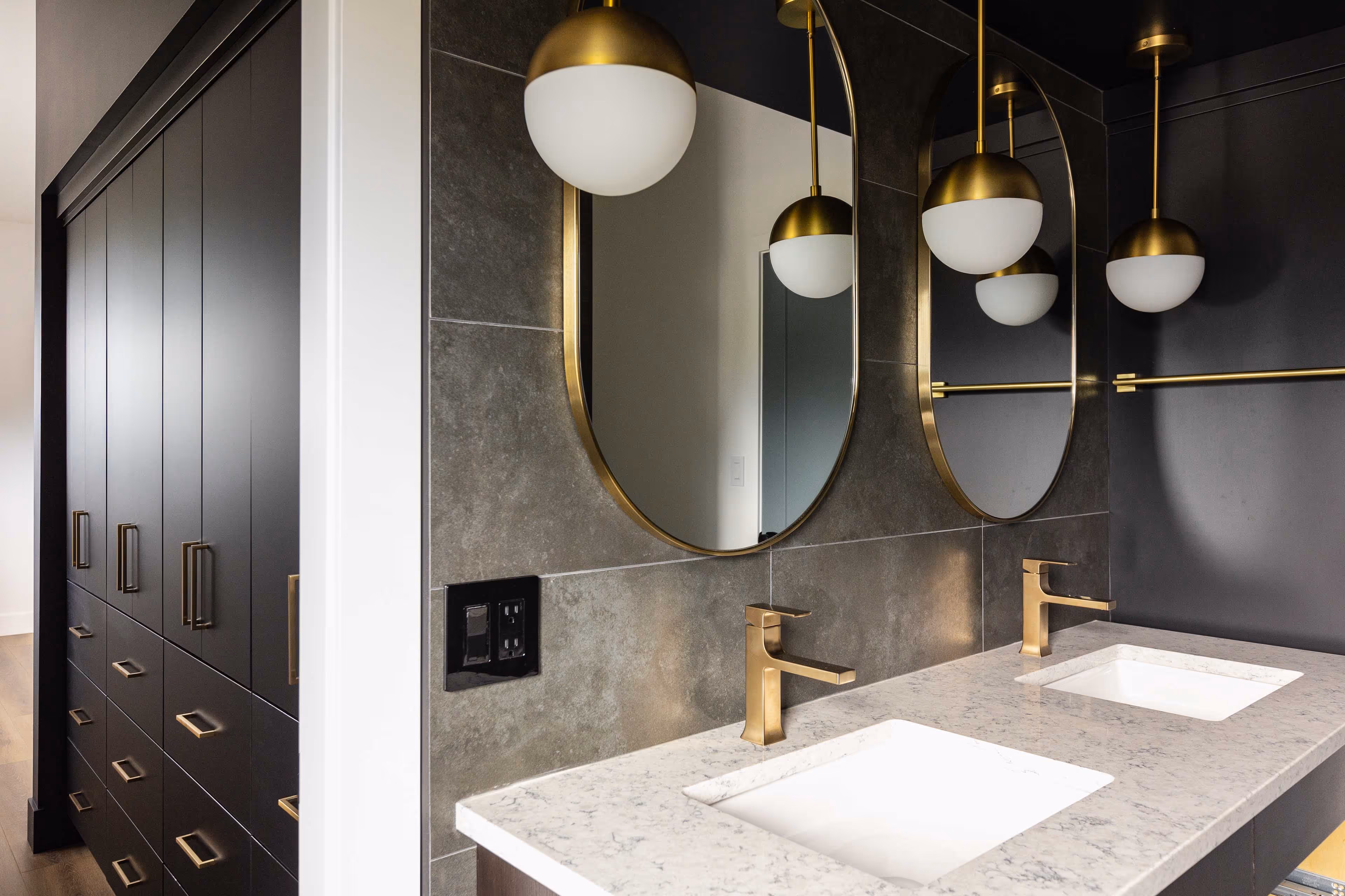 Modern bathroom with double white sinks, gold faucets, oval mirrors with gold frames, and dark cabinetry with gold handles.