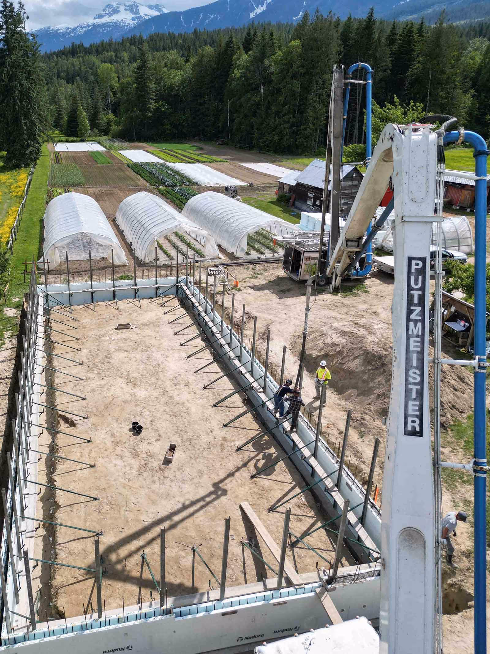 Construction site with concrete wall framework and a large Putzmeister pump crane on a farm with greenhouses and mountain backdrop.