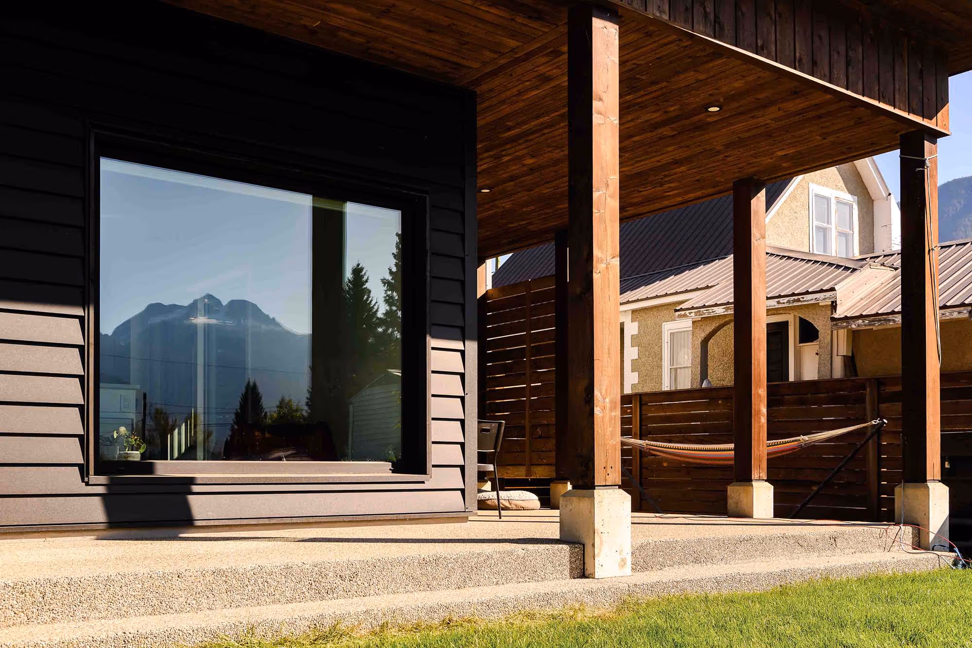 Covered wooden deck with concrete base, chair, and hammock, next to a house with a large window reflecting a mountain and trees.