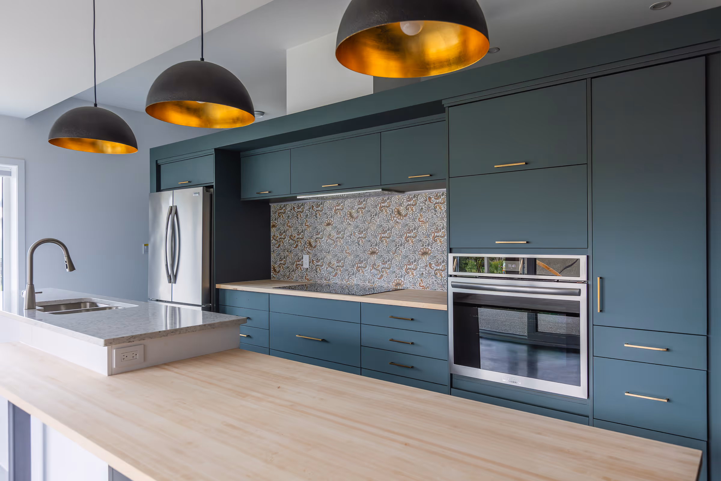 Modern kitchen with teal cabinetry, patterned tile backsplash, wooden countertops, stainless steel appliances, and three black and gold pendant lights.
