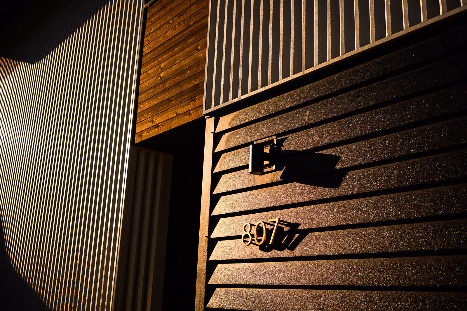 Sunlit modern exterior wall with horizontal siding, house number 807, and a mounted light casting shadows.