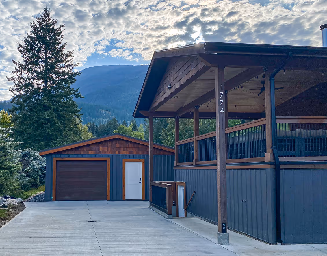 Modern detached garage with wood and dark blue paneling, adjacent to a house with a large covered porch, surrounded by trees and mountains under a partly cloudy sky.