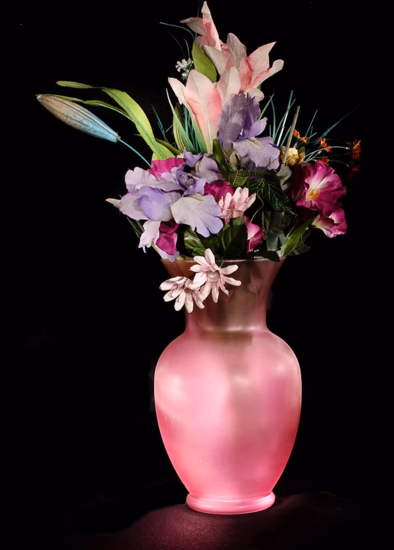Pink frosted glass vase holding an assortment of colorful artificial flowers on a black background.