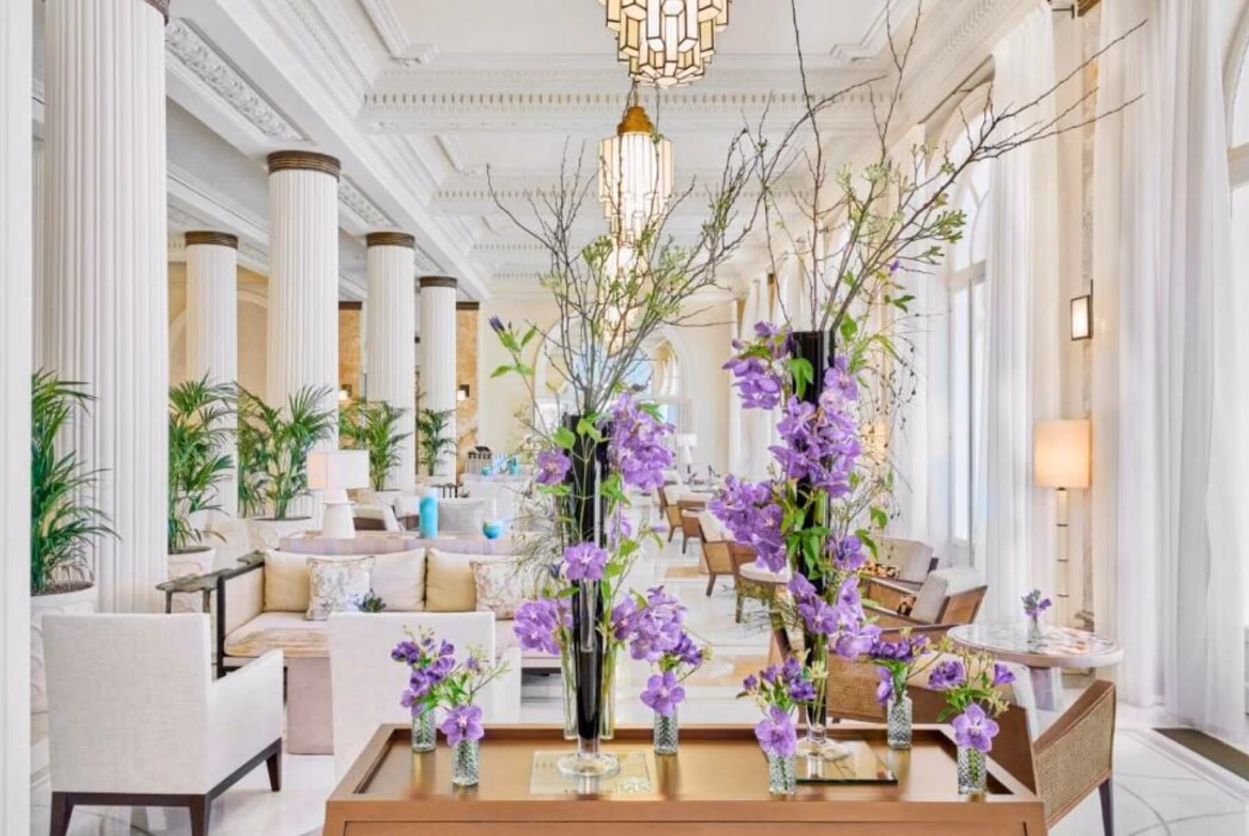 Elegant hotel lobby with white pillars, beige seating, large windows, and purple floral arrangements.