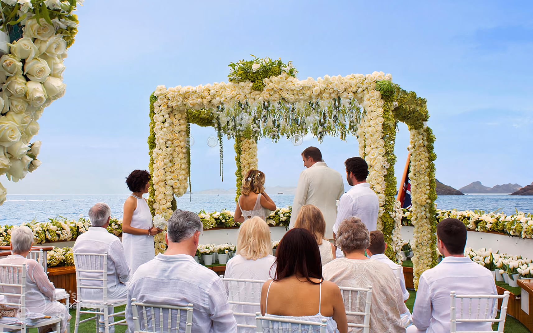 Wedding ceremony with bride and groom under a floral arch by the ocean, guests seated in white chairs.