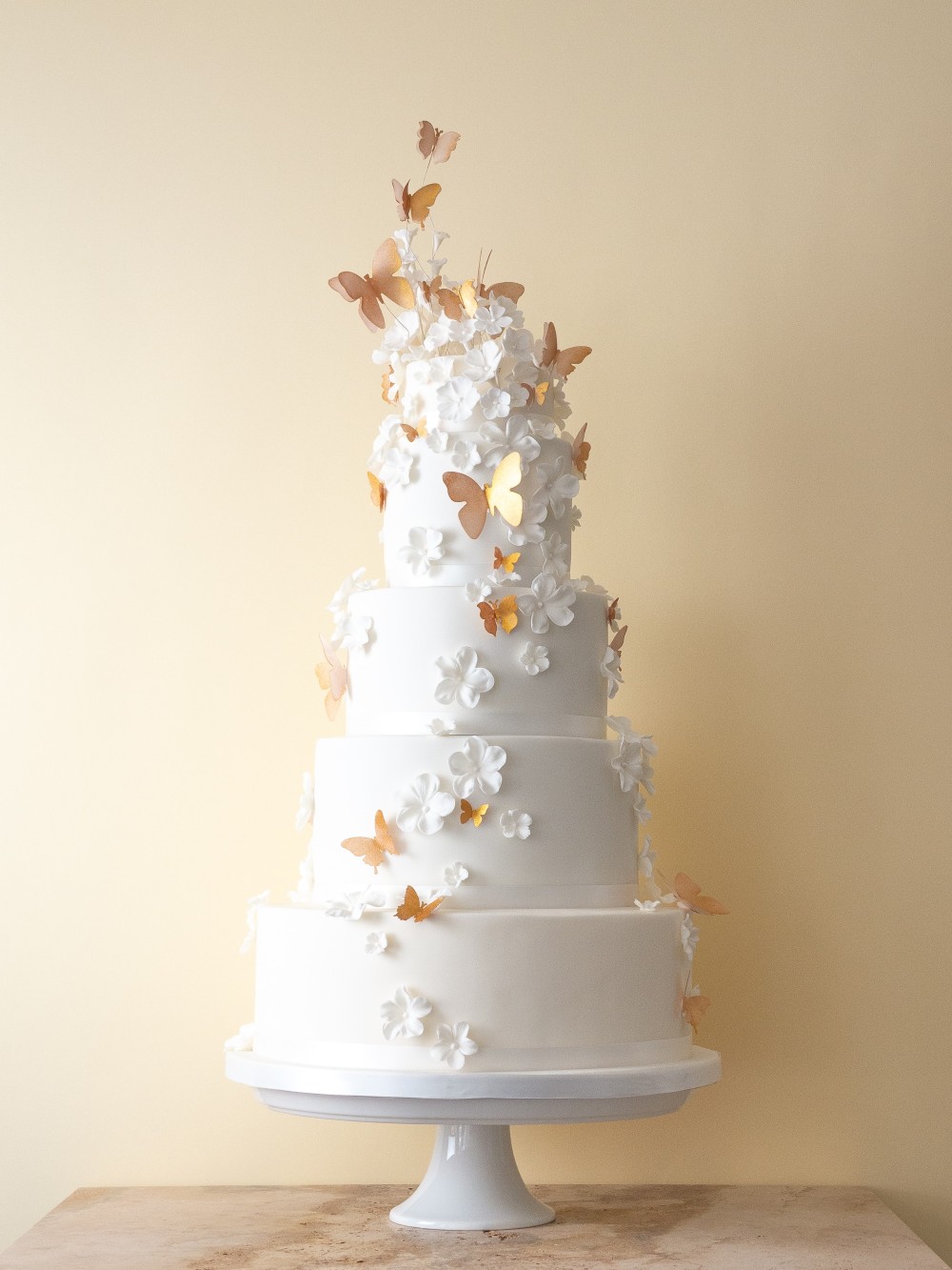 Four tier white wedding cake with cascading white blossoms and edible wafer paper butterflies in warm golden tones.