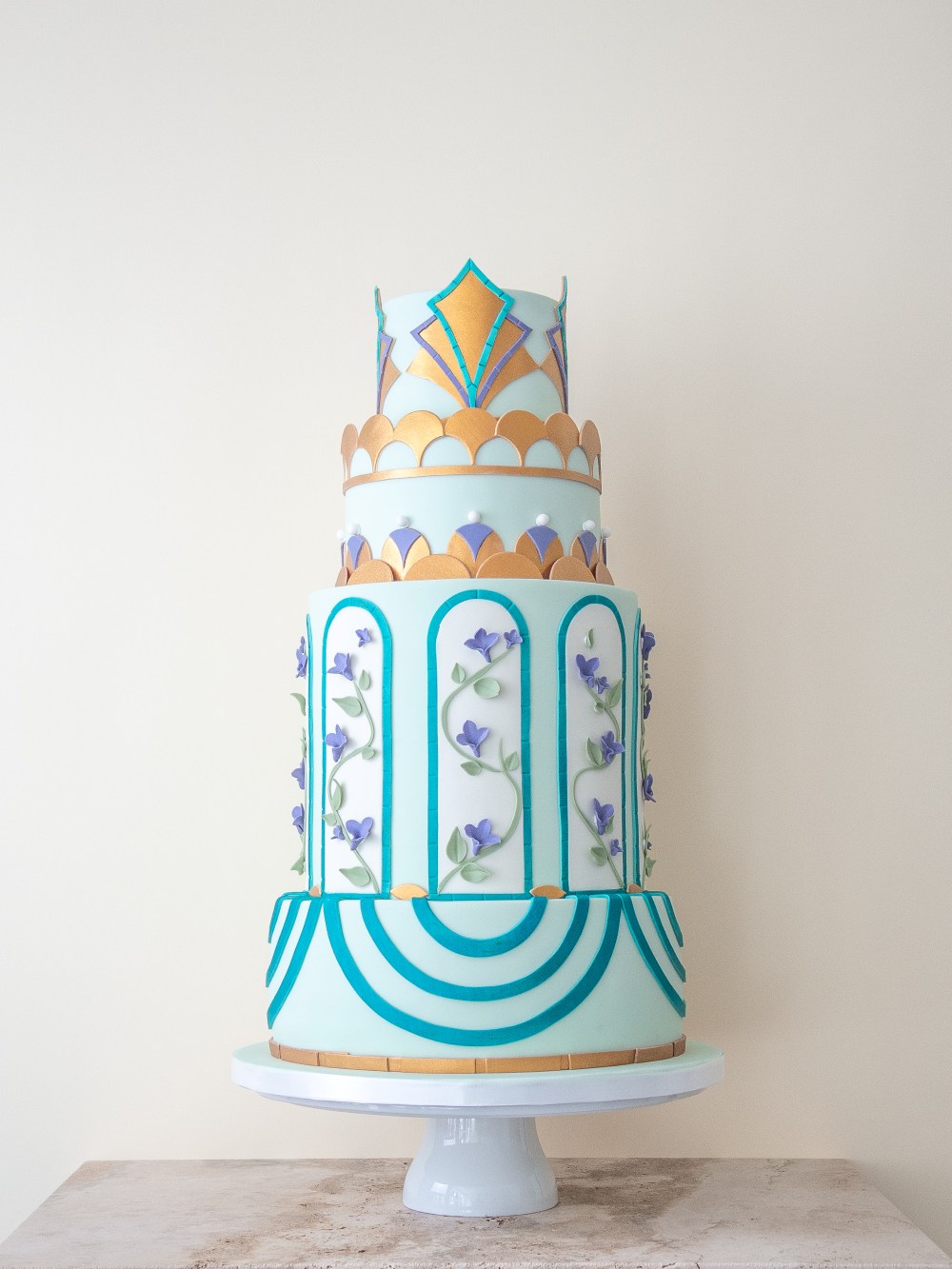 Four tier wedding cake in powder blue with teal Art Deco arches, gold geometric detailing, and lilac floral vines in arched panels.