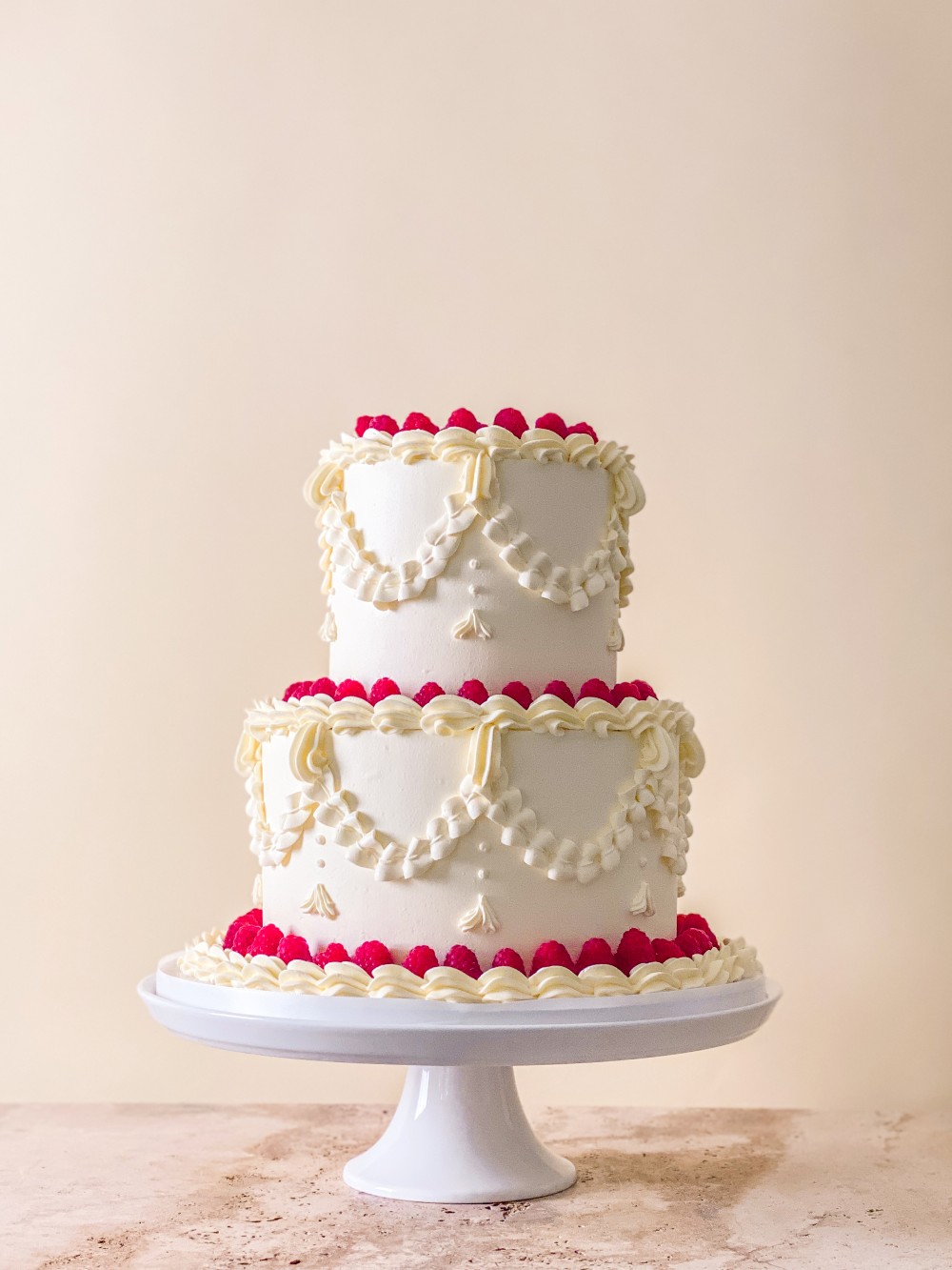 Two tier white wedding cake with piped buttercream swags and borders, topped and finished with fresh raspberries.