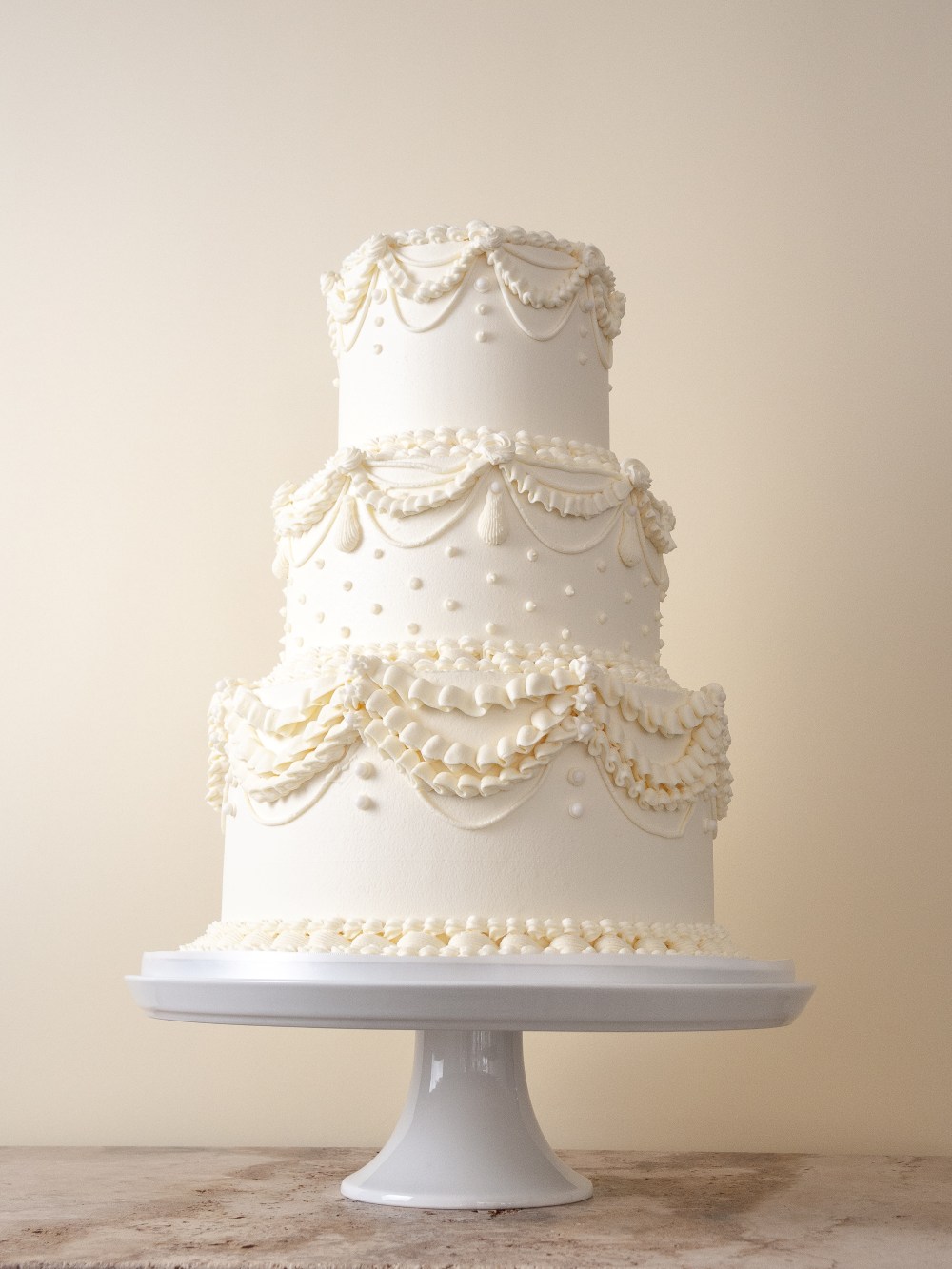 Three tier ivory wedding cake with baroque style piped swags, ruffles, pearl dots, and ornate buttercream detailing.