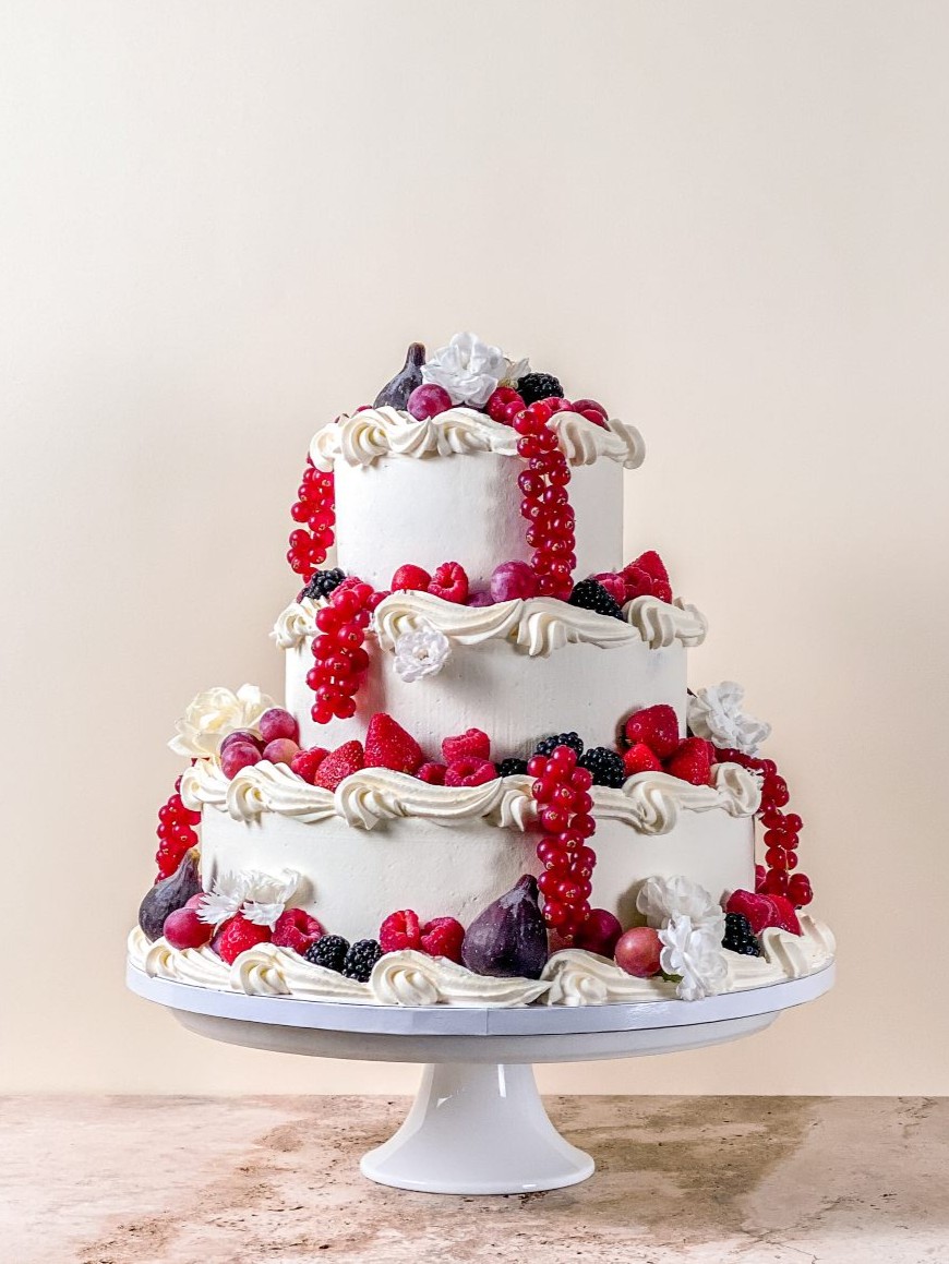 Three tier white celebration cake with piped vanilla swirls, topped with fresh berries, figs and cascading redcurrants, finished with white blossoms.