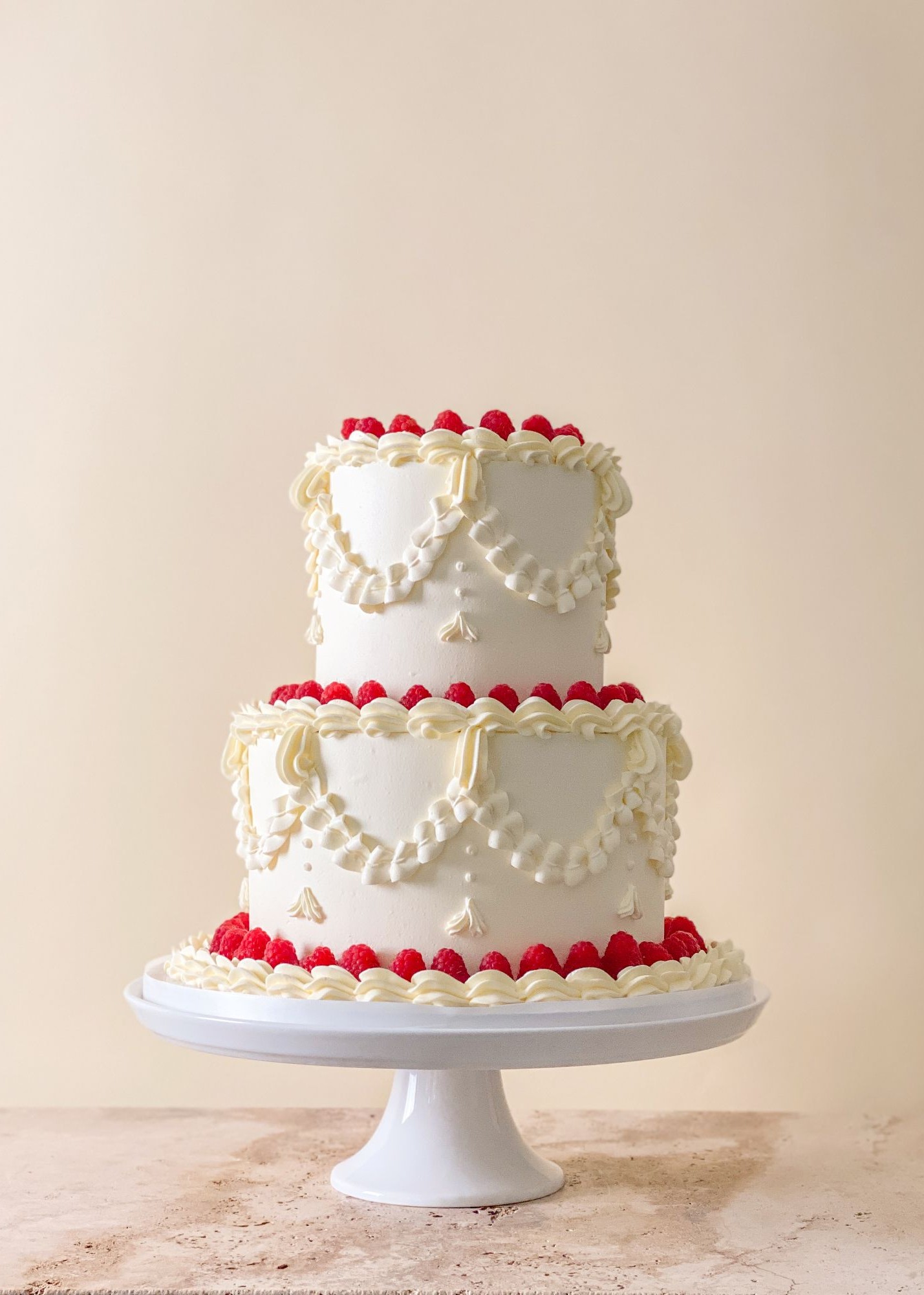 Two tier white wedding cake with piped buttercream swags and borders, topped and finished with fresh raspberries.