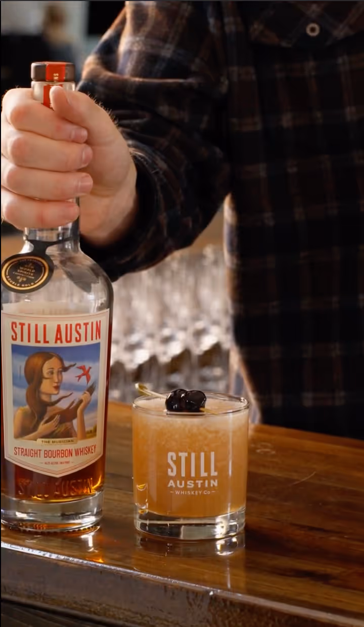 Hand holding a bottle of Still Austin Straight Bourbon Whiskey next to a whiskey glass with cherries on a wooden bar counter.